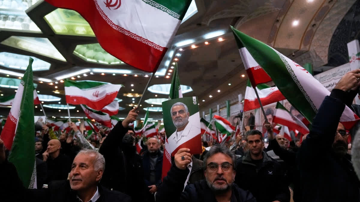 People wave Iranian flags as one of them holds up a poster of the late commander of the Iran's Revolutionary Guard expeditionary Quds Force, Gen. Qassem Soleimani, who was killed in a U.S. drone attack in 2020 in Iraq, during a ceremony commemorating his death anniversary at the Imam Khomeini grand mosque in Tehran, Iran, Thursday, Jan. 1, 2026.