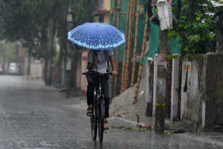 A commuter on a cycle holds an umbrella while passing through a rain-slicked road amid rainfall, in Siliguri.