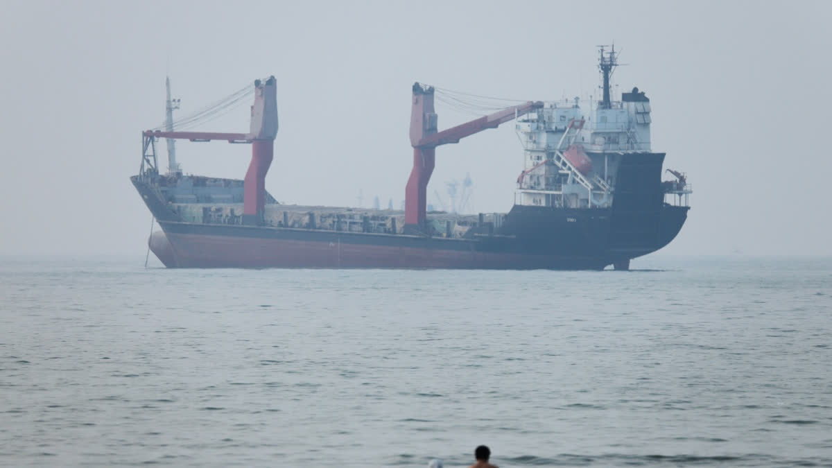 A group of people stands in shallow water as a cargo ship appears anchored in the Strait of Hormuz off Bandar Abbas, Iran