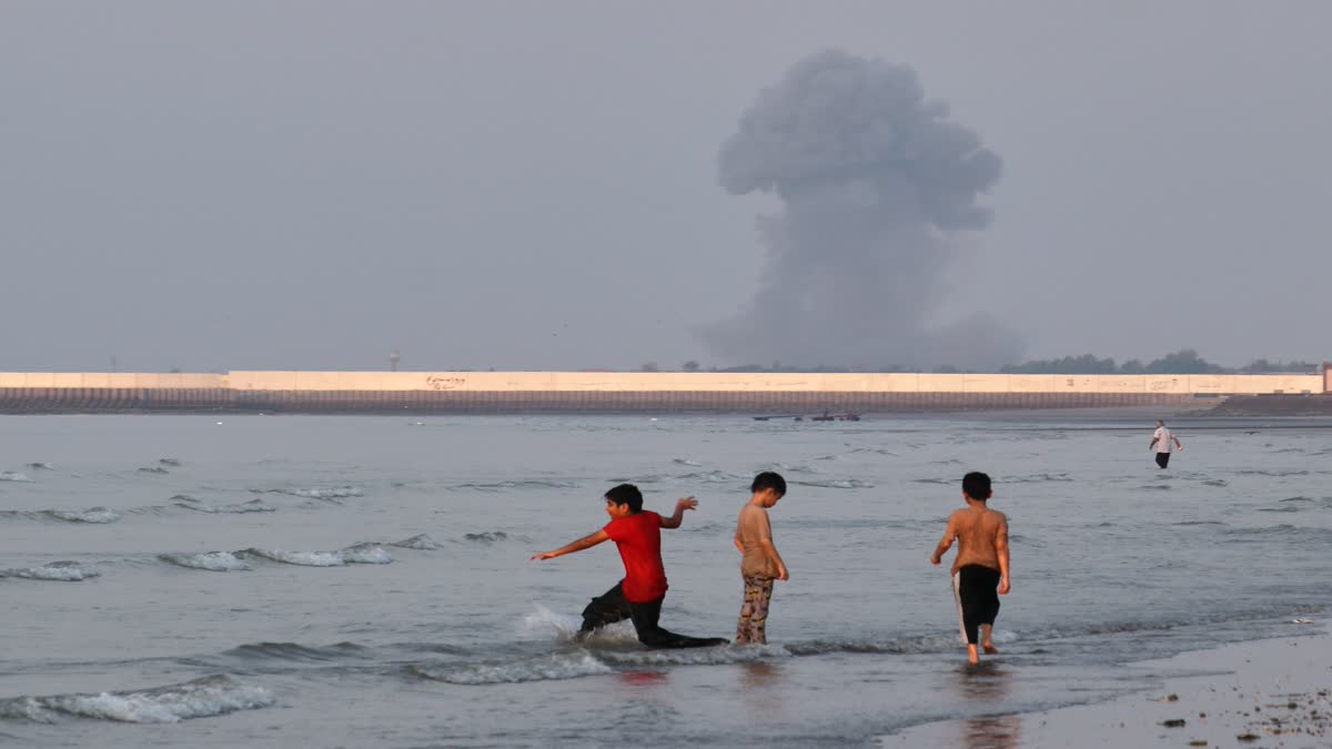 Three boys play in the shallow waters of the Strait of Hormuz, as a plume of smoke rises from an explosion in the background, off Bandar Abbas, Iran, Monday,