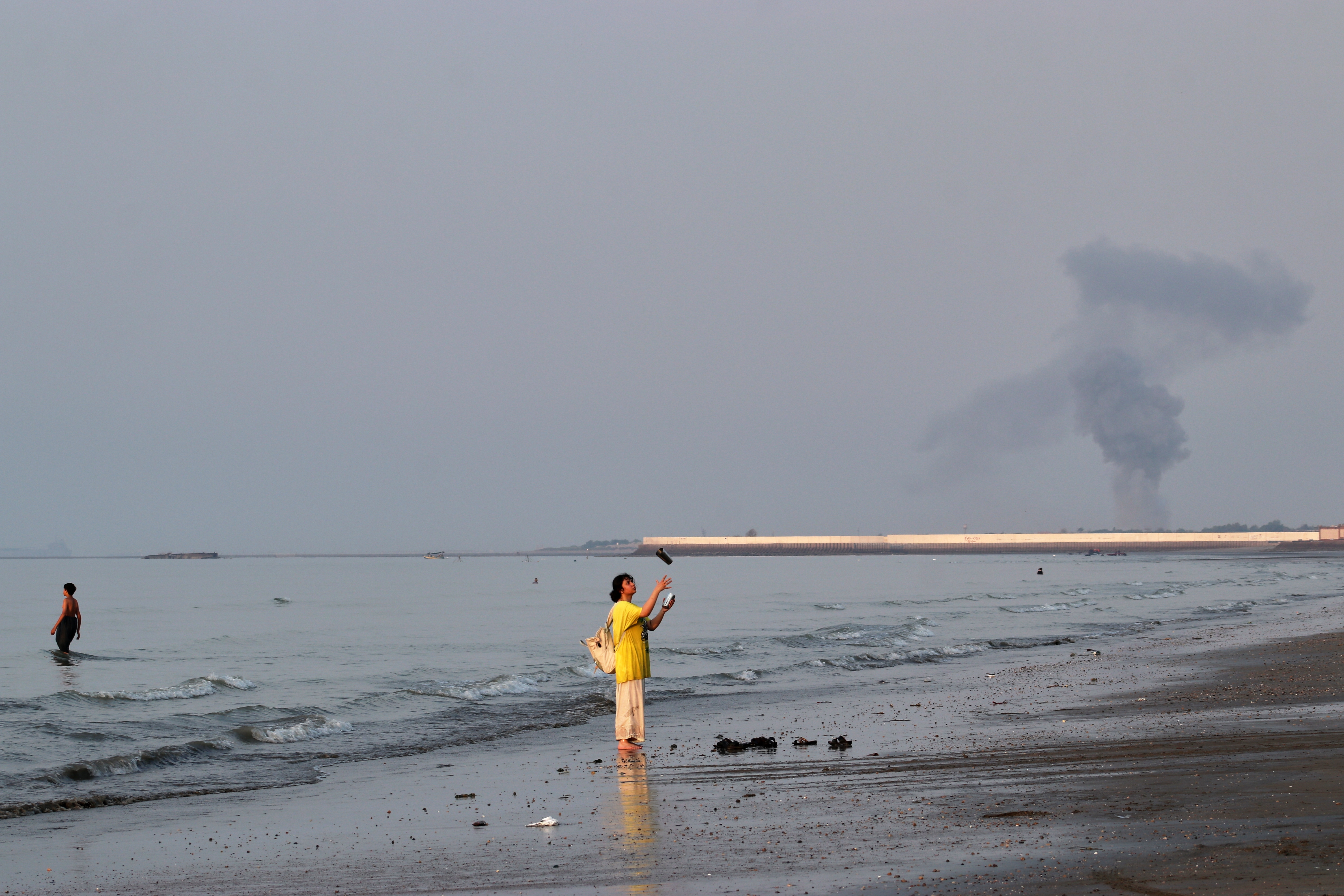 A woman stands at the water's edge along the Strait of Hormuz as a plume of smoke rises in the background following an explosion, off Bandar Abbas, Iran, Monday, July 13, 2026.
